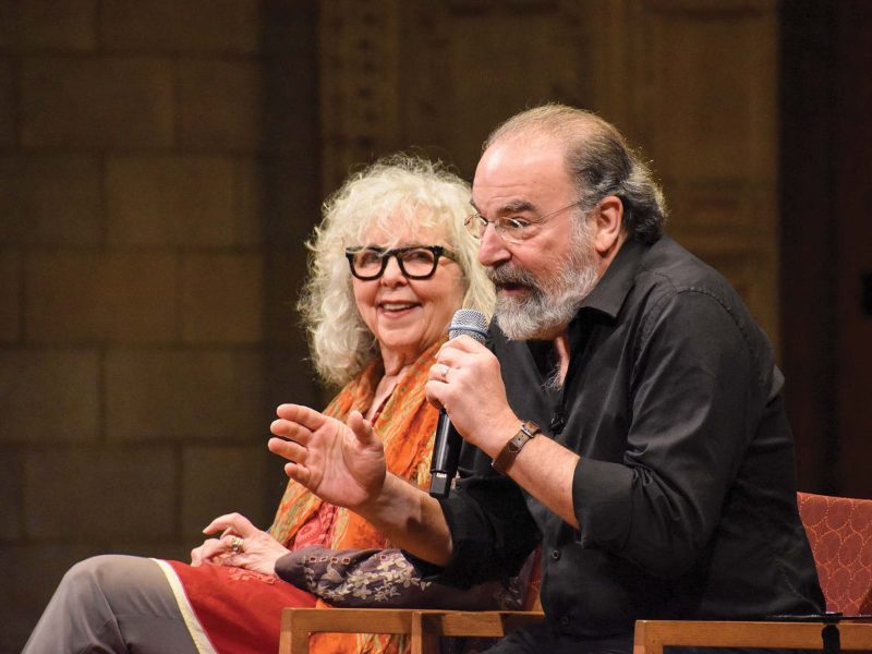 A woman and man, both gray-haired and bespectacled, seated. The man leans forward to speak into a handheld microphone.