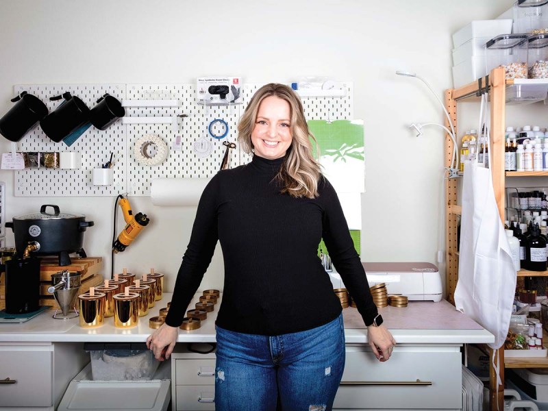 A blonde woman in a turtleneck and jeans standing in front of a desk. The desk, wall behind her and shelves to her side contain tools and materials, some of which are parts of candles.