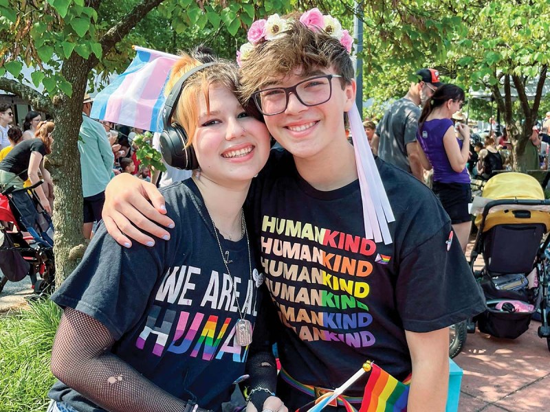 Two young people standing, one with arm around the other, wearing Pride-related and rainbow apparel and accessories