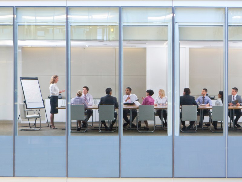 many people in business attire seated at a conference table listening to one speak, seen through the windows of their building