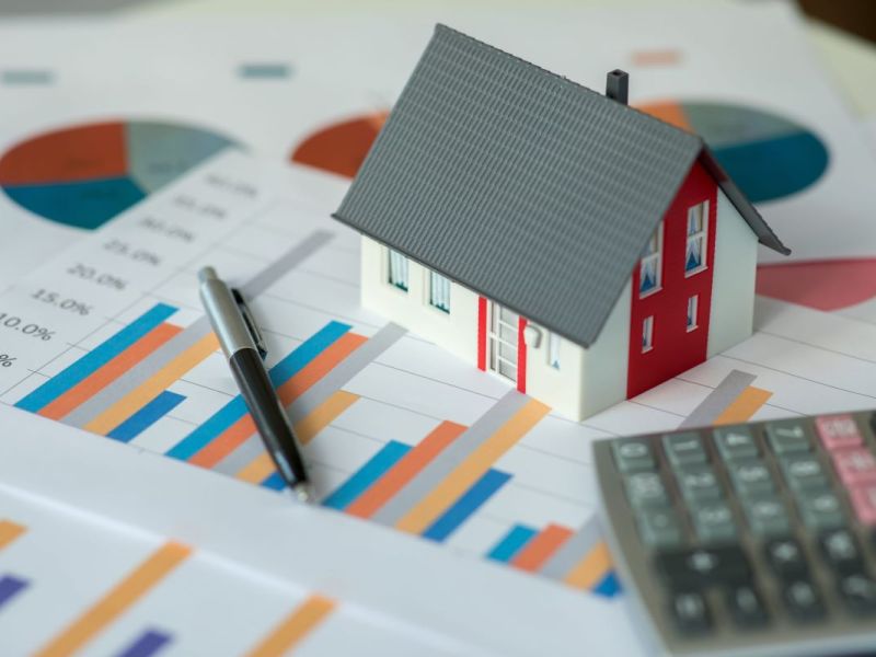 A model of a house, a pen, and a calculator are propped on top of stacks of paper graphs.