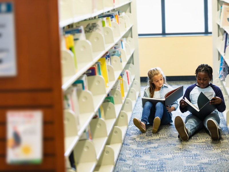 Two children reading books on the floor of a library.