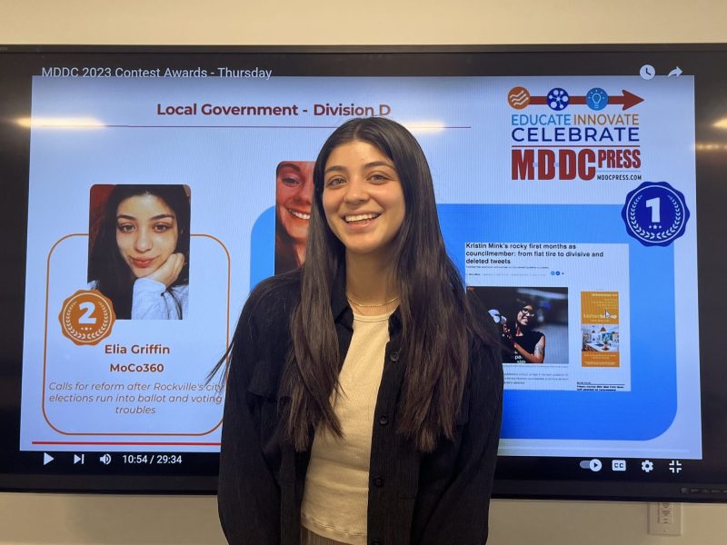 A smiling young woman- it's Elia- stands in front of a tv screen displaying the winners of a Local Government award. She got second prize!