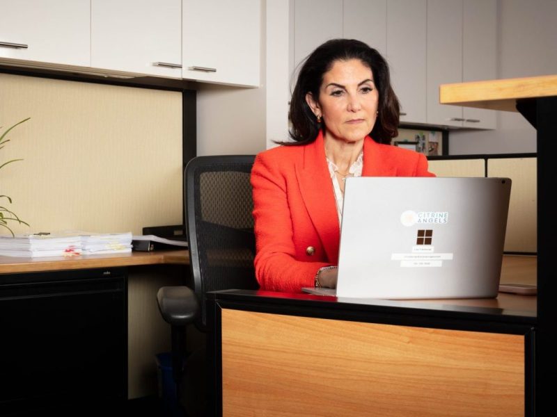 A woman in a red blazer sits at a desk with a laptop