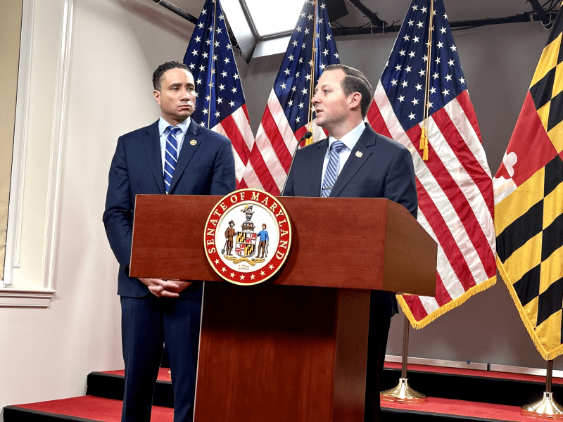 two men at a podium, american and maryland flags behind them